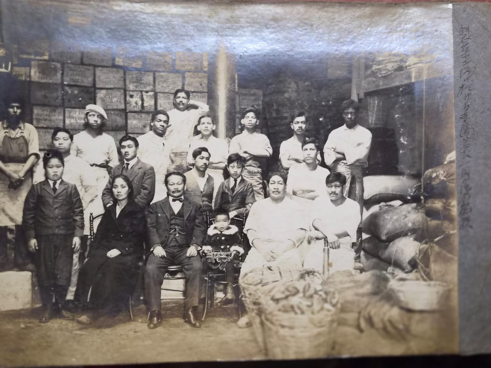 Grupo de hombres, mujeres y niños fotografiados en el interior de un almacén, rodeados de sacos, cestas y mercadería, en una fotografía histórica vinculada a la comunidad japonesa en el Perú.