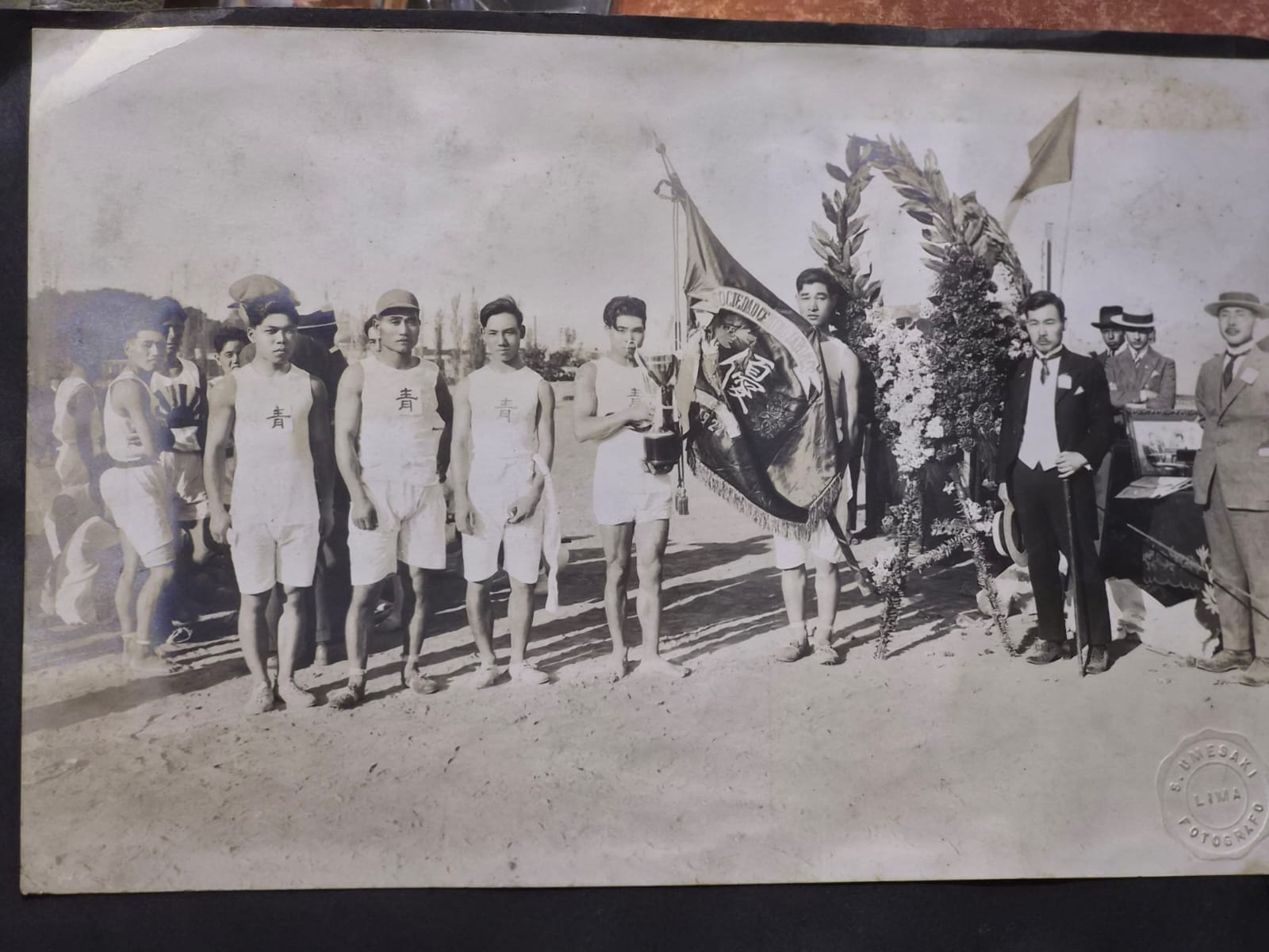 Fotografía histórica de jóvenes deportistas con uniformes blancos, acompañados de un trofeo, un estandarte, un arreglo floral y varios hombres vestidos de traje en Lima.