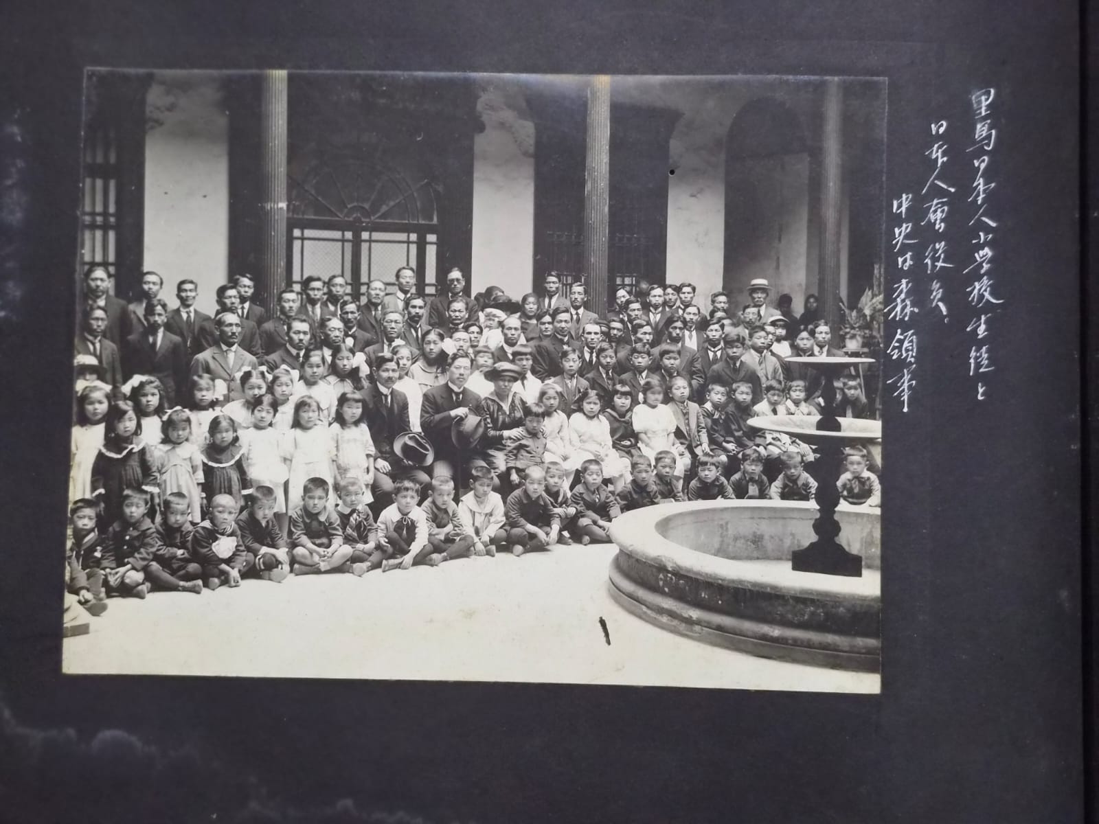 Fotografía histórica de un numeroso grupo de adultos y niños reunidos en el patio de un edificio con columnas y una fuente, con inscripciones manuscritas en japonés.